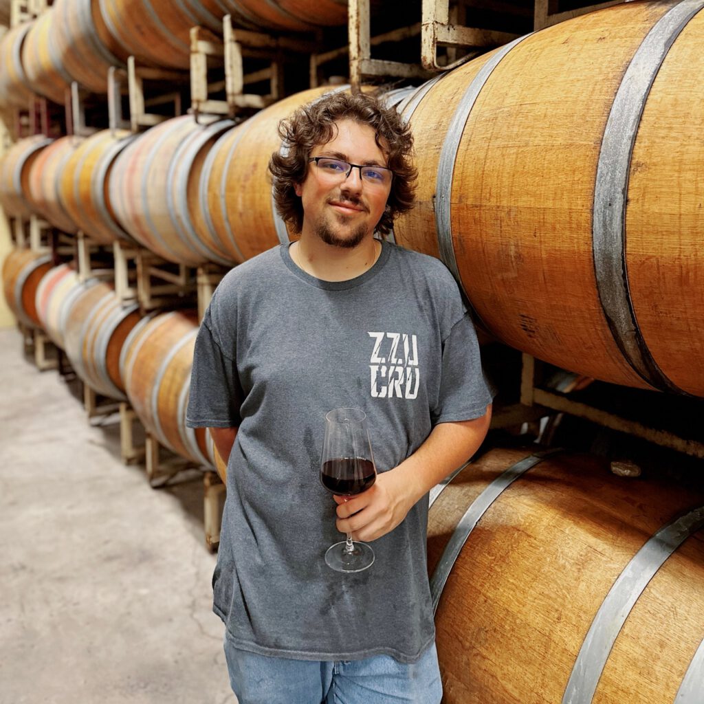 Assistant Winemaker Jared Grace with a wine glass leaning against barrels. 
